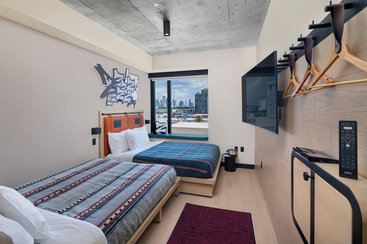 A modern hotel room with two beds, a large window with a city view, a mounted TV, and a coat rack.