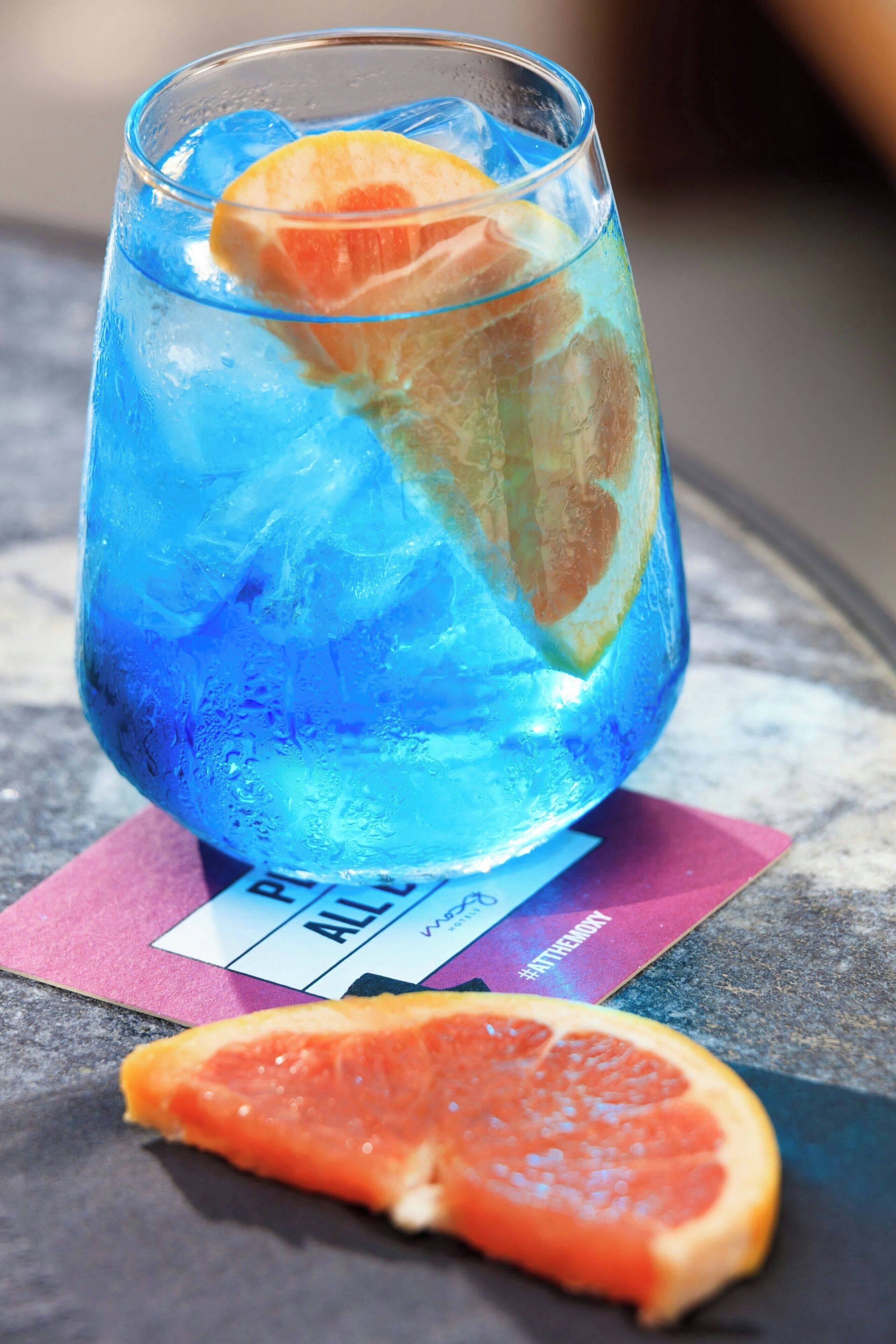 A glass with blue liquid and an orange slice, placed on a table with a pink napkin.