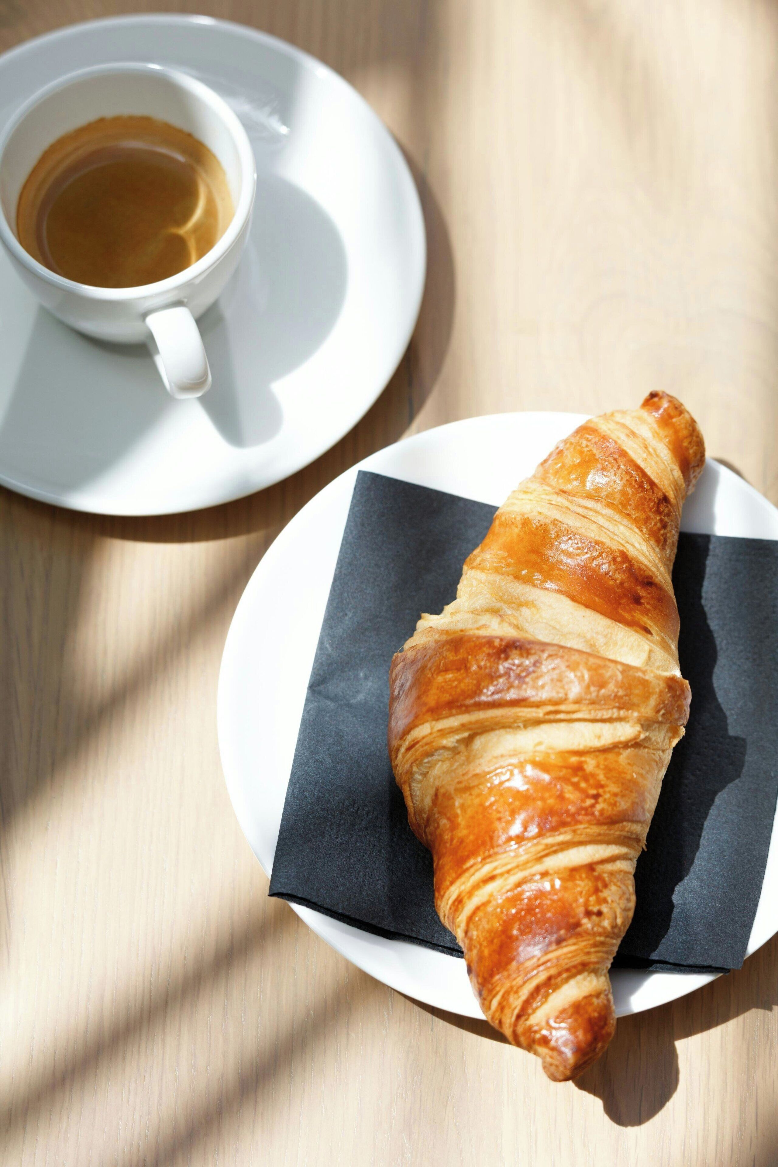 En croissant på en tallrik med en servett, och en kopp kaffe på ett litet fat bredvid.