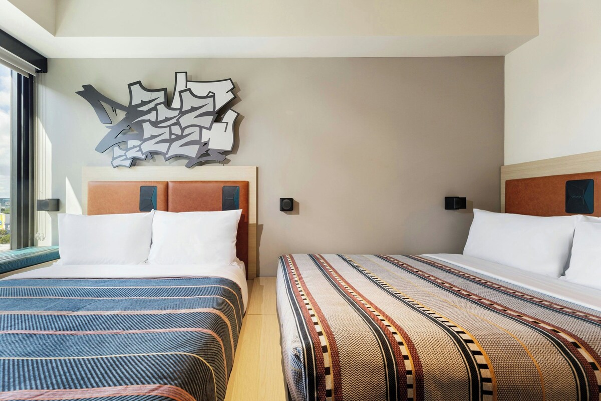 A hotel room with two beds, a modern abstract wall art, and a large window providing a view.