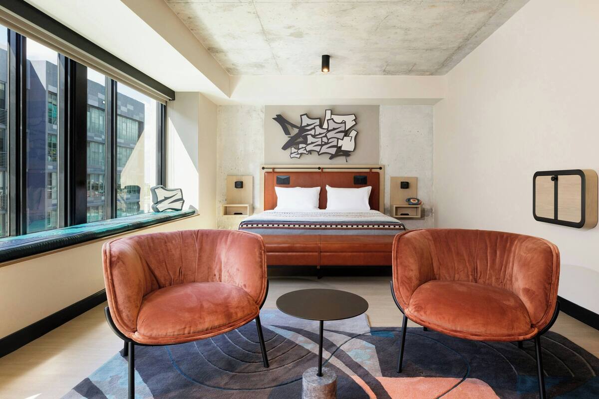 A modern hotel room with a large bed, two leather chairs, a small table, and a city view through large windows.