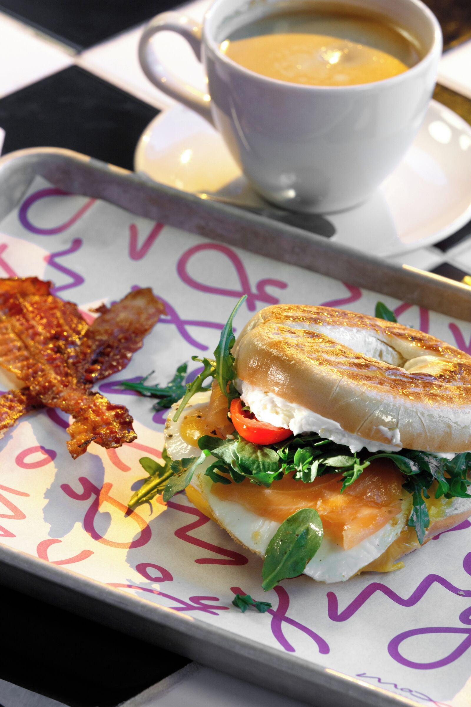 A breakfast tray with a bagel sandwich, bacon, and a cup of coffee.