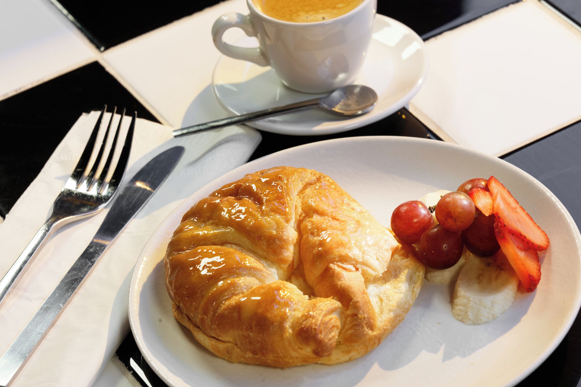 A croissant, strawberries, and a banana on a plate with a cup of coffee and a spoon on a table.