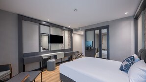 A modern hotel room with a large bed, a desk with a flat-screen TV, a seating area with a chair and a small table, and a large mirror.