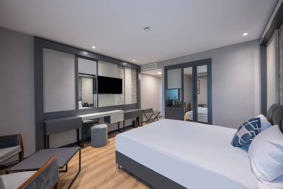 Une chambre d’hôtel moderne dotée d’un grand lit, d’un bureau avec un téléviseur à écran plat, d’un coin salon avec un fauteuil et une petite table, ainsi que d’un grand miroir.