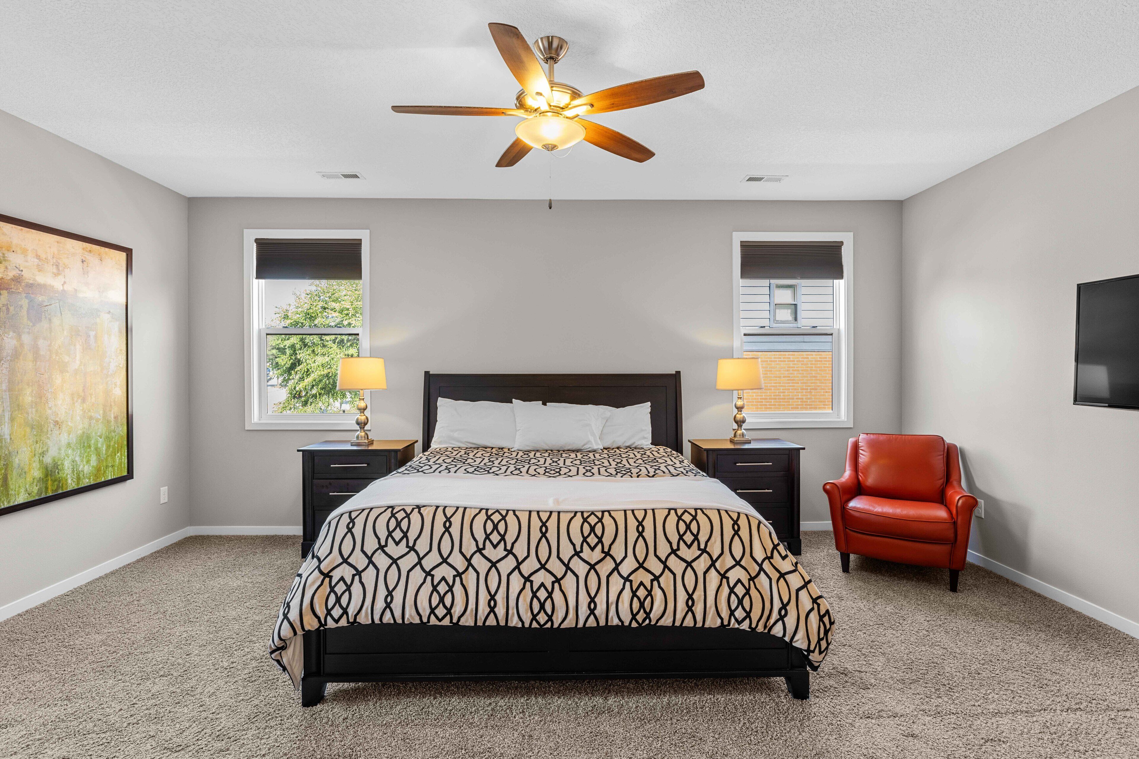 A bedroom with a bed, a ceiling fan, two bedside lamps, a red chair, and a large painting on the wall.