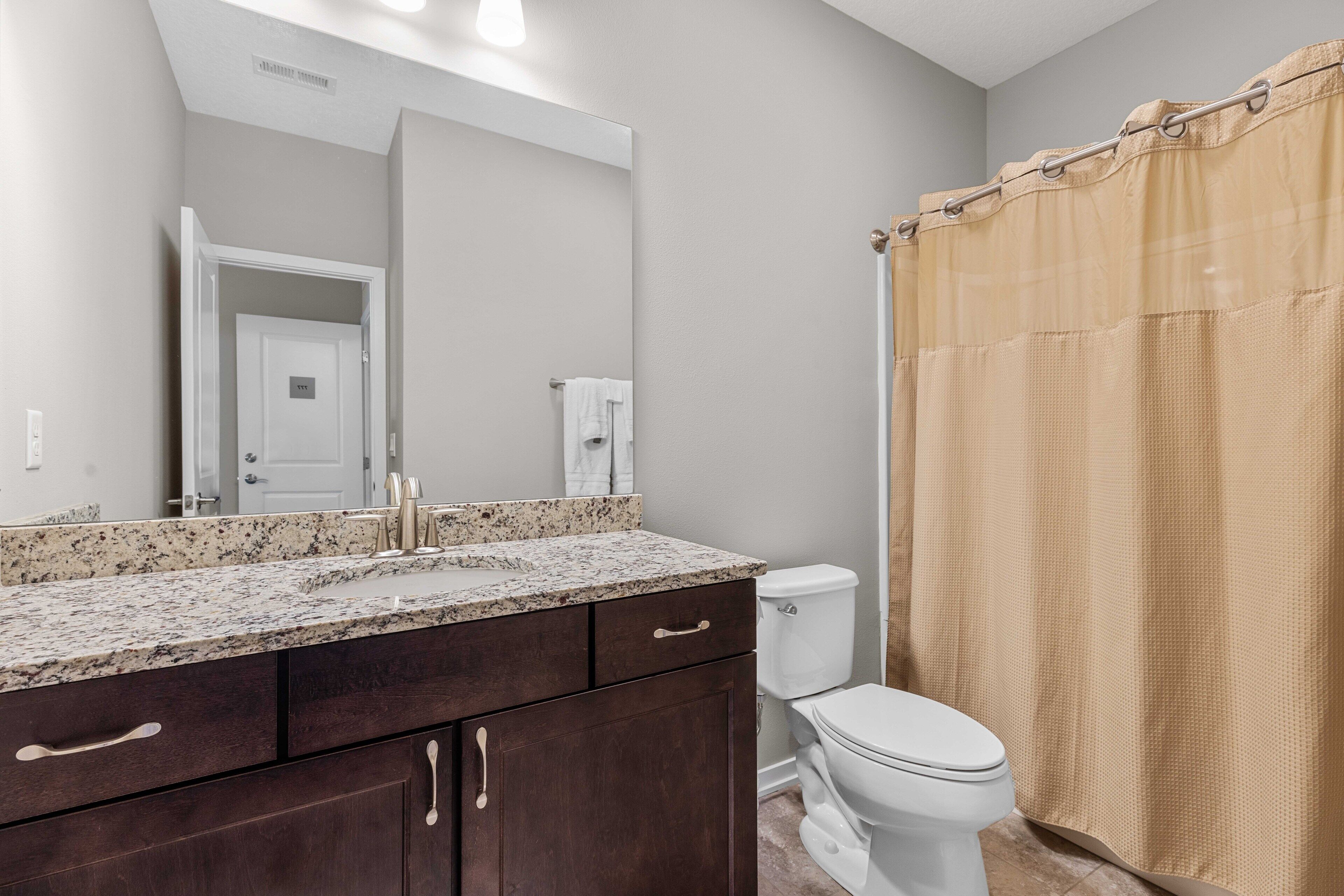A bathroom with a granite countertop sink, dark wooden cabinets, a white toilet, and a beige shower curtain.