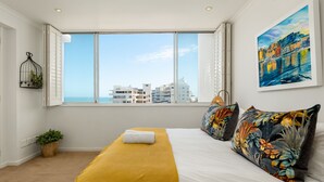 A bedroom with a large window, a bed with white linens, a yellow throw blanket, a tropical-themed pillow, and a cityscape view. - Stunning Bantry Bay Haven - Beach on Your Doorstep (Cape Town)
