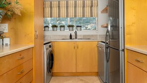 A compact kitchen with wooden cabinets, a stainless steel refrigerator, a washing machine, and a window with potted plants. - Stunning Bantry Bay Haven - Beach on Your Doorstep (Cape Town)