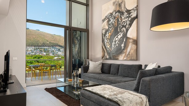 A modern living room with a large window, a grey sofa, a glass coffee table, and a painting on the wall. - Lux Elegance in Heart of Mouille Point (Cape Town)