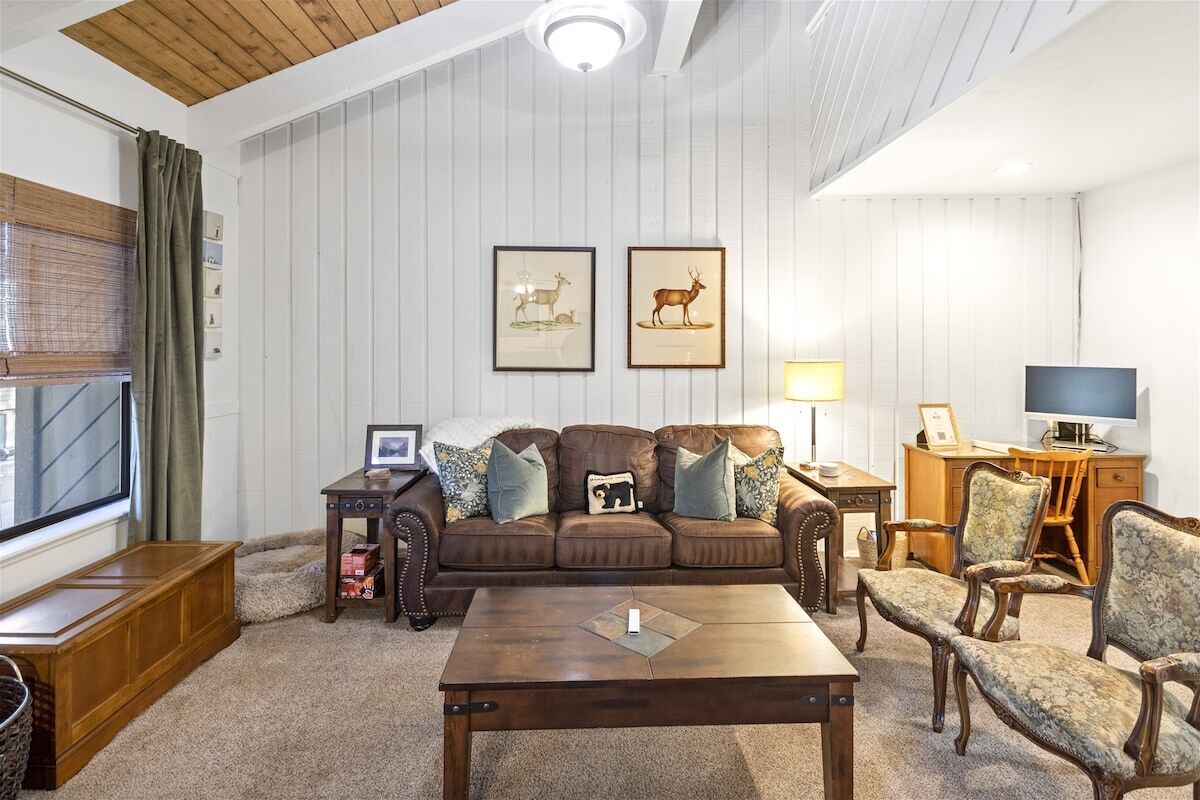 Un salon chaleureux avec un canapé en cuir marron, une table basse en bois et un téléviseur posé sur un meuble en bois. Des tableaux encadrés ornent le mur, et une fenêtre est agrémentée de rideaux.
