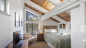 A bedroom with a sloped ceiling, a bed, a TV, a chair, and a window with blinds. - Sunshine Village 126: Pet friendly, Close to Green (Mammoth Lakes)