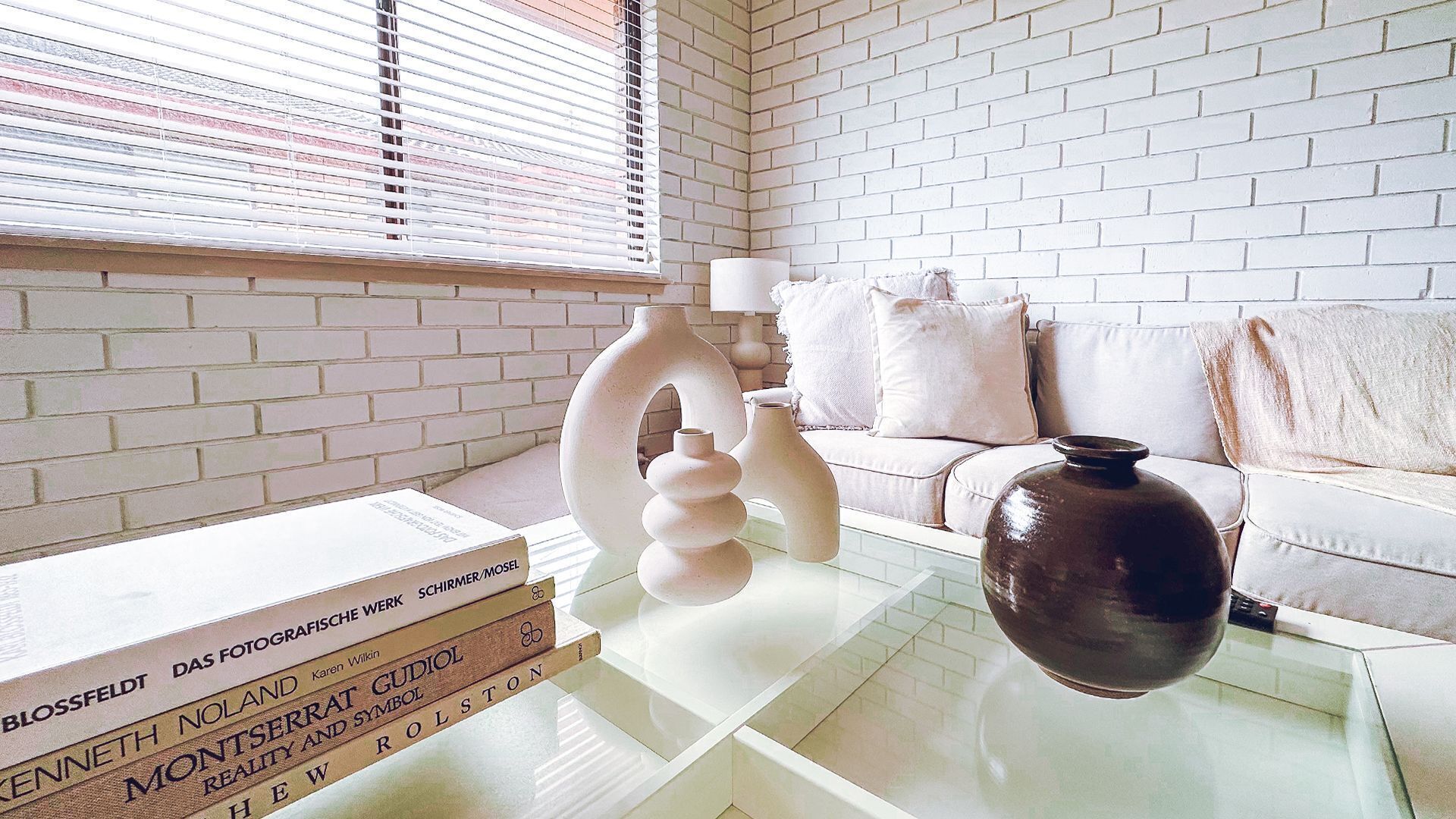 Una sala de estar moderna con una pared de ladrillo blanco, una mesa de centro de vidrio, un jarrón marrón y un sofá blanco con cojines.