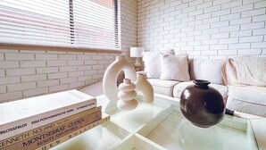 Ein modernes Wohnzimmer mit weißer Ziegelwand, einem Glastisch, einer braunen Vase und einem weißen Sofa mit Kissen.