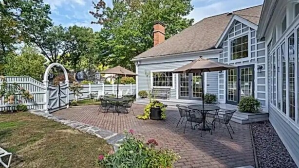 Un patio con mobiliario exterior, una zona de estar cubierta y un sendero de ladrillo que conduce a una casa con amplias ventanas y chimenea.
