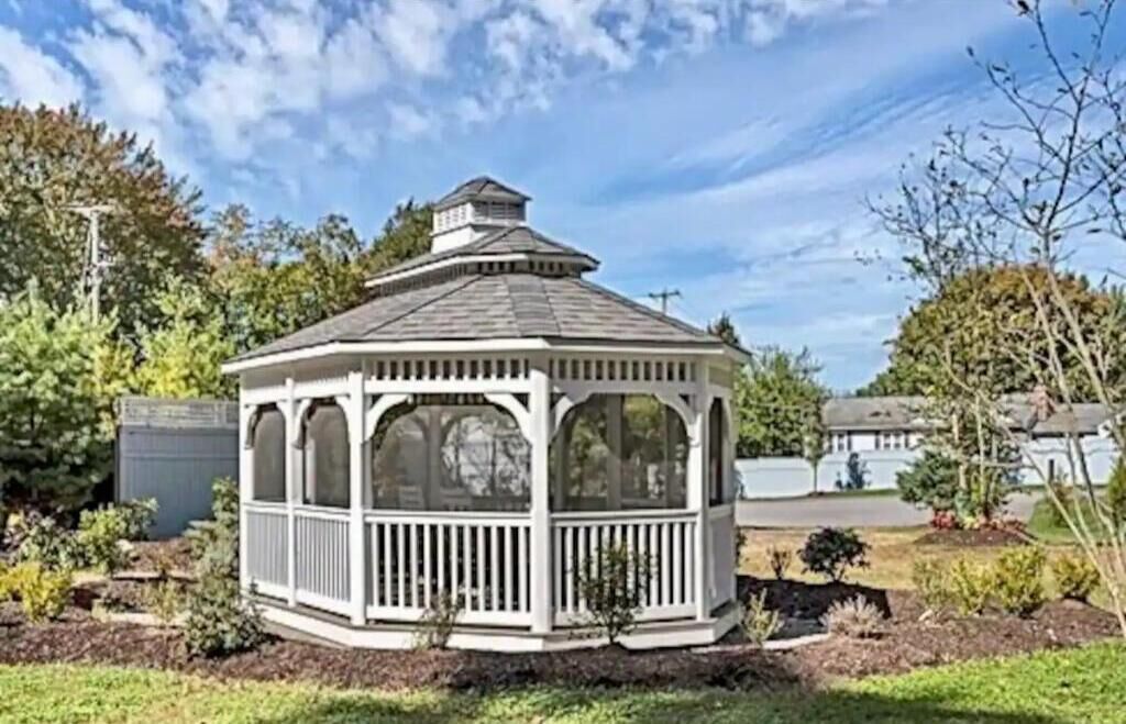 A white gazebo with a peaked roof and a balcony, surrounded by a garden with various plants and a lawn.