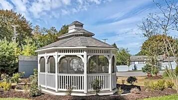 A white gazebo with a peaked roof and a balcony, surrounded by a garden with various plants and a lawn.