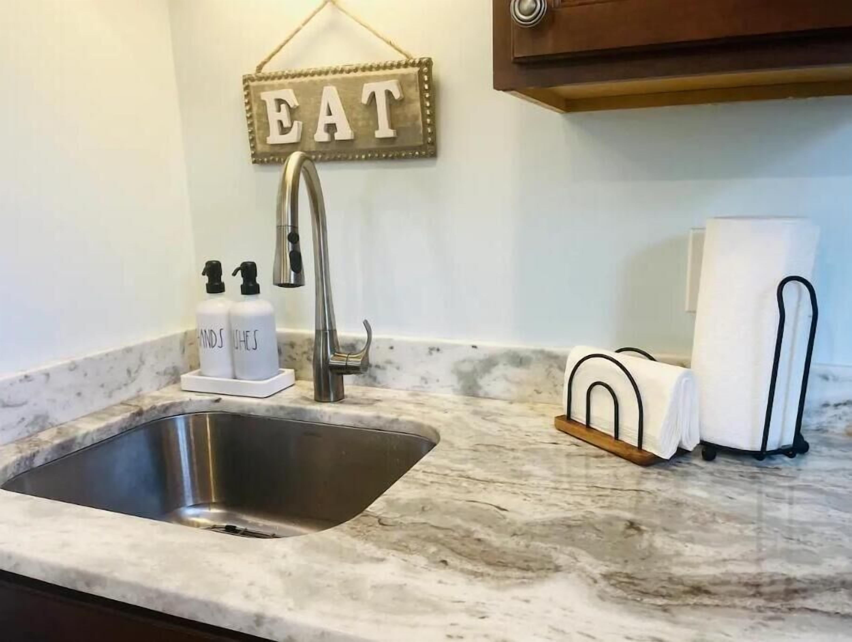 Una cocina con un lavamanos de acero inoxidable, encimera de mármol, grifo, dispensadores de jabón y un perchero para toallas.