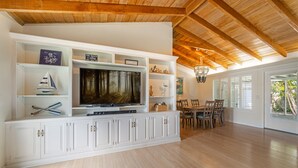 A spacious living room with a large flat-screen TV mounted on a white cabinet, wooden flooring, and a dining area with a wooden table and chairs.