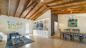 A spacious living room with a high ceiling, wooden beams, a dining area with a table and chairs, and a large window with blinds.