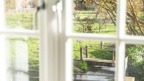 Blick durch ein Fenster auf eine ländliche Landschaft mit einer kleinen Brücke über einem Bach, einem weißen Zaun und einem Gebäude mit einem schrägen Dach.