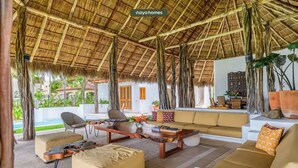 A spacious living area with a thatched roof, comfortable seating, and a view of a pool.