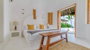 A modern bedroom with a bed, bedside tables, a wooden bench, and a view of palm trees through an open door.