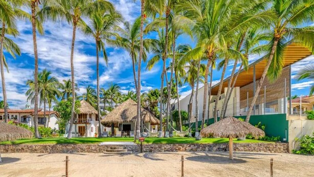A resort with thatched-roof huts, palm trees, and a modern building.