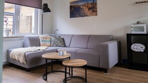 A living room with a grey sofa, two round wooden tables, a beach-themed pillow, a beach-themed bag, a picture of a beach, a window with a view of a building, and a shelf with a microwave and a rolled-up towel.