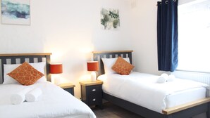 Habitación de hotel con dos camas, cada una con sábanas blancas y almohadones marrones estampados. Hay mesitas de noche con lámparas y una ventana con cortinas azules.