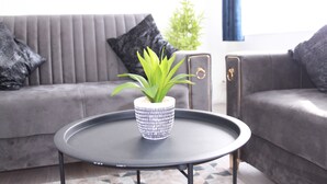 Una sala moderna con un sofá gris, una mesita redonda con una planta en maceta, y una planta decorativa al fondo.