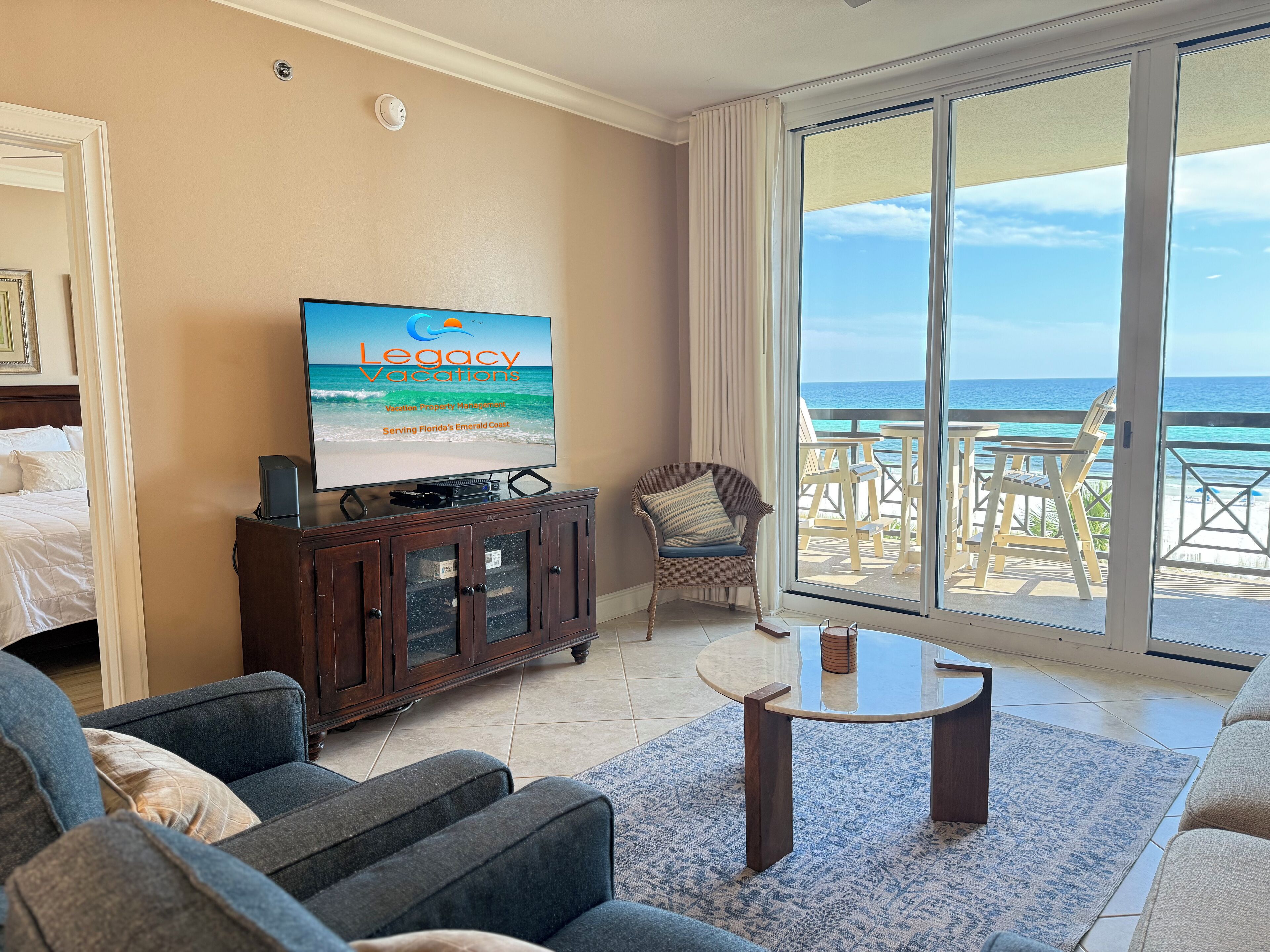 A hotel room with a television displaying a vacation property management service, a balcony with a view of the ocean, and a seating area with a table.