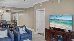 A living room with a television displaying 'Legacy Vacations' and a dining area with chairs and a table. - Azure 404 | 4BD  | Gulf Views + Covered Balcony | FREE Beach Setup (Fort Walton Beach)