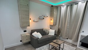 A modern living room with a grey sofa, a wooden coffee table, and a white rug. There is a wooden cabinet and a small bedside table with a lamp. - Charmant Appartement Joinvillais (Joinville-le-Pont)
