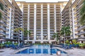 A multi-story hotel with a central swimming pool surrounded by lounge chairs and palm trees.
