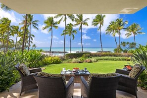 Un patio con mobiliario de mimbre y una mesa lista para el desayuno, con vistas a una playa con palmeras y un cielo azul despejado.