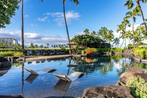 A clear pool with lounge chairs and palm trees, surrounded by a landscaped area with a view of a hotel building. - Luxury Ko Olina Beach Villa B109 by Gather w/ Stunning Ocean & Kapolei Views (Kapolei)