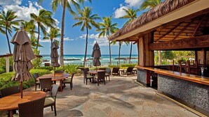 A patio area with tables and chairs, a bar, and a view of the ocean with palm trees.