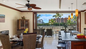A dining area with wicker chairs, a central island with a fruit bowl, and a view of palm trees and buildings through large windows.