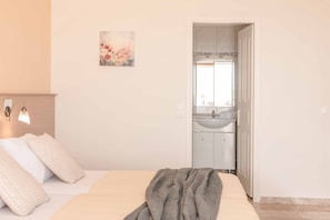 Ein Schlafzimmer mit Bett, Kissen, Decke, einem Gemälde und Blick auf ein Badezimmer mit Waschbecken und Spiegel.