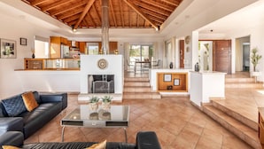 A spacious living area with a high wooden ceiling, a fireplace, a kitchen with modern appliances, and a dining area.