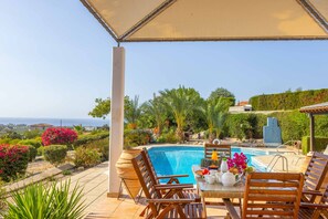 Zona junto a la piscina con vista al mar, rodeada de exuberante vegetación y una mesa lista para comer.