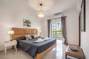 Een slaapkamer met een bed, nachtkastjes, een hanglamp, een raam met gordijnen en uitzicht op een balkon.