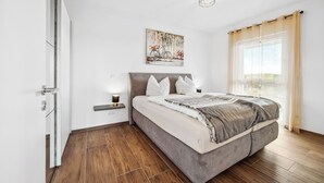 Un dormitorio moderno con cama, mesita de noche, una obra de arte en la pared y ventana con cortinas.