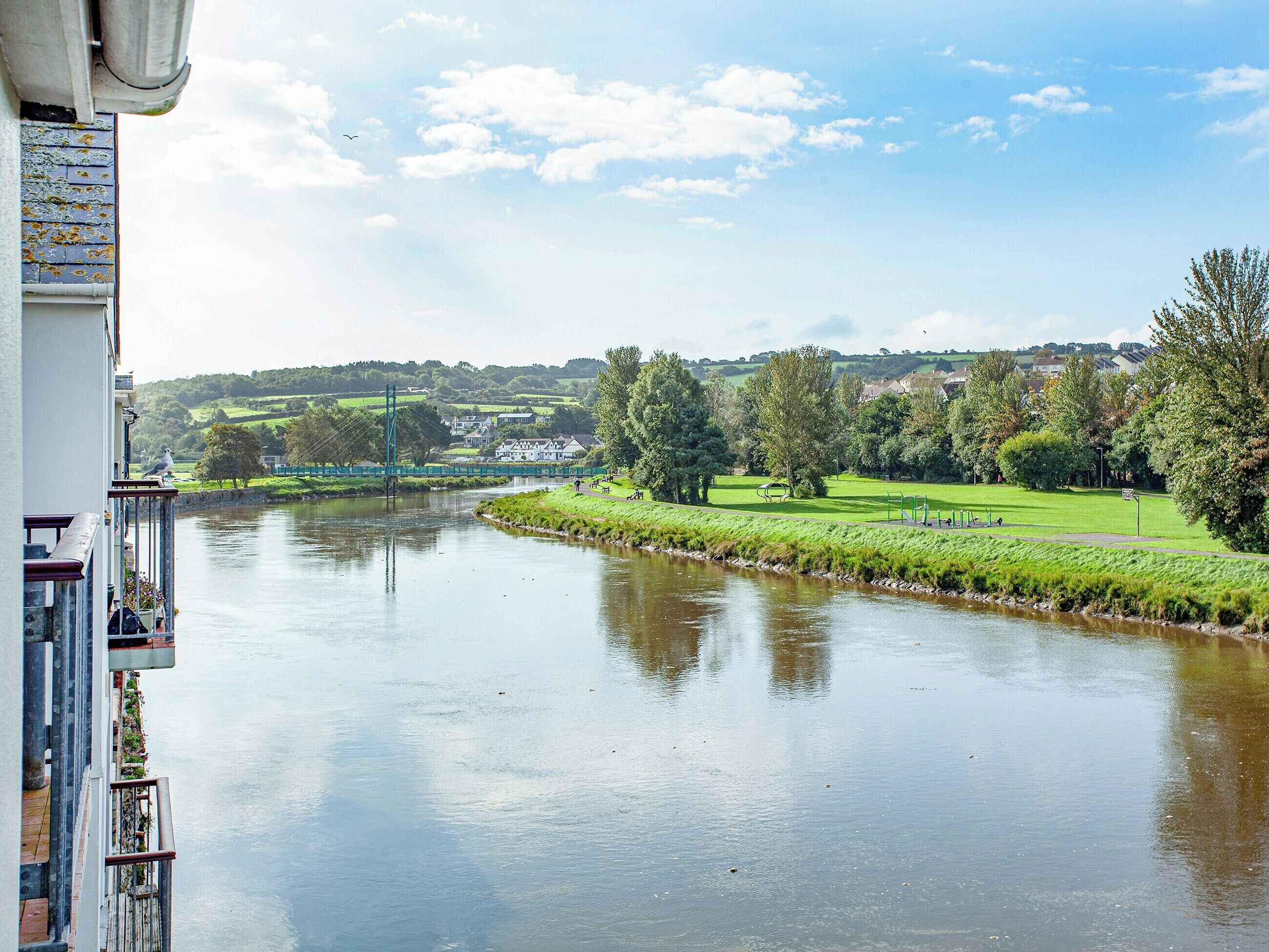 Ein Fluss mit einer Brücke, Grünflächen und Häusern im Hintergrund.
