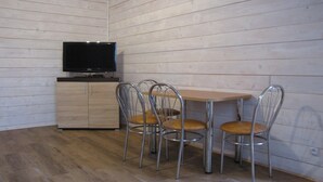 Een knus ingerichte kamer met een houten wand, een flatscreen televisie op een houten kast en een kleine eettafel met vier metalen stoelen.