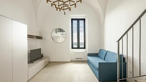 Una sala de estar moderna con un televisor sobre un mueble blanco, un sofá azul, un espejo redondo y una ventana con vistas.