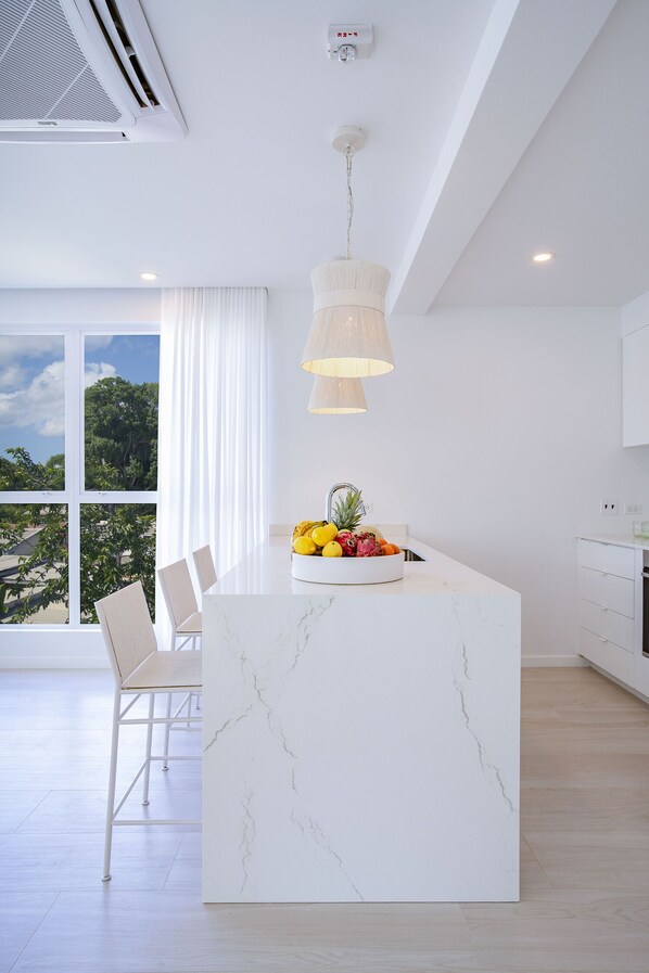 A modern kitchen with a marble island, white cabinetry, and a large window with a view of trees. - Palm Grove Villas & Hotel (Mount Standfast)