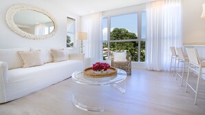 A spacious living room with a white sofa, a round mirror, a clear coffee table with a woven tray, and a view of the outdoors through large windows. - Palm Grove Villas & Hotel (Mount Standfast)