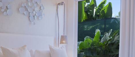 A bedroom with a large bed, a ceiling fan, a wall-mounted light, and a view of greenery through a window.